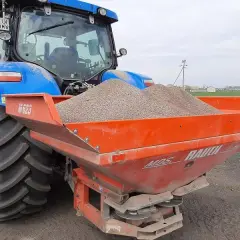 Niebieski traktor z przyczepionym rozsiewaczem napełnionym materiałem do odkażania gleby wapnem.