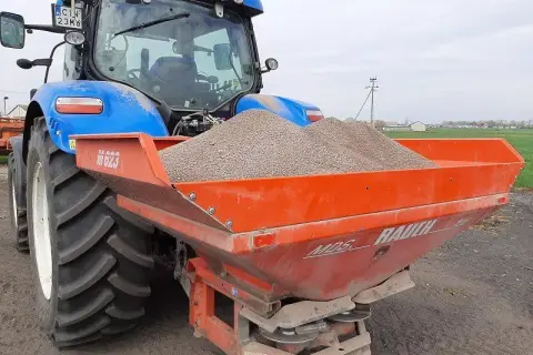 Niebieski traktor z przyczepionym rozsiewaczem napełnionym materiałem do odkażania gleby wapnem.