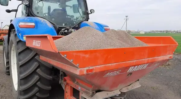 Niebieski traktor z przyczepionym rozsiewaczem napełnionym materiałem do odkażania gleby wapnem.