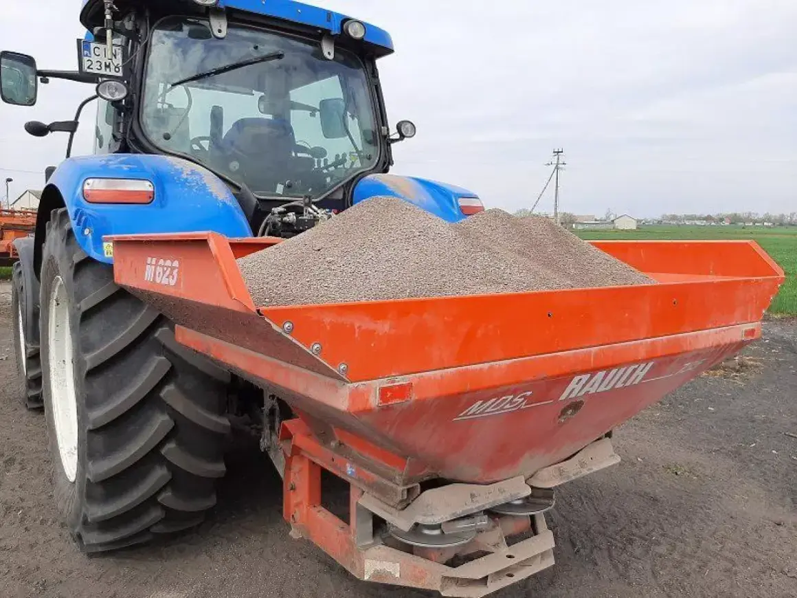 Niebieski traktor z przyczepionym rozsiewaczem napełnionym materiałem do odkażania gleby wapnem.